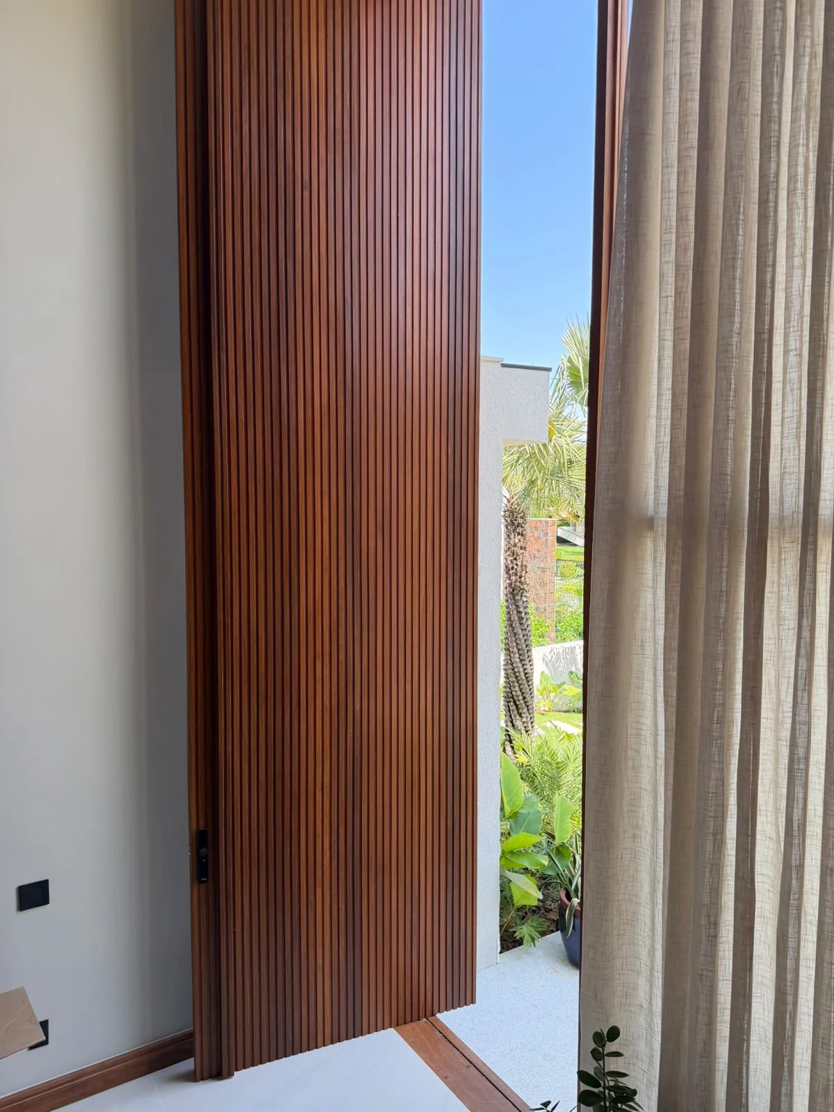 Porta de madeira com frisos verticais entreaberta, com ferragem escura e vista para o jardim.